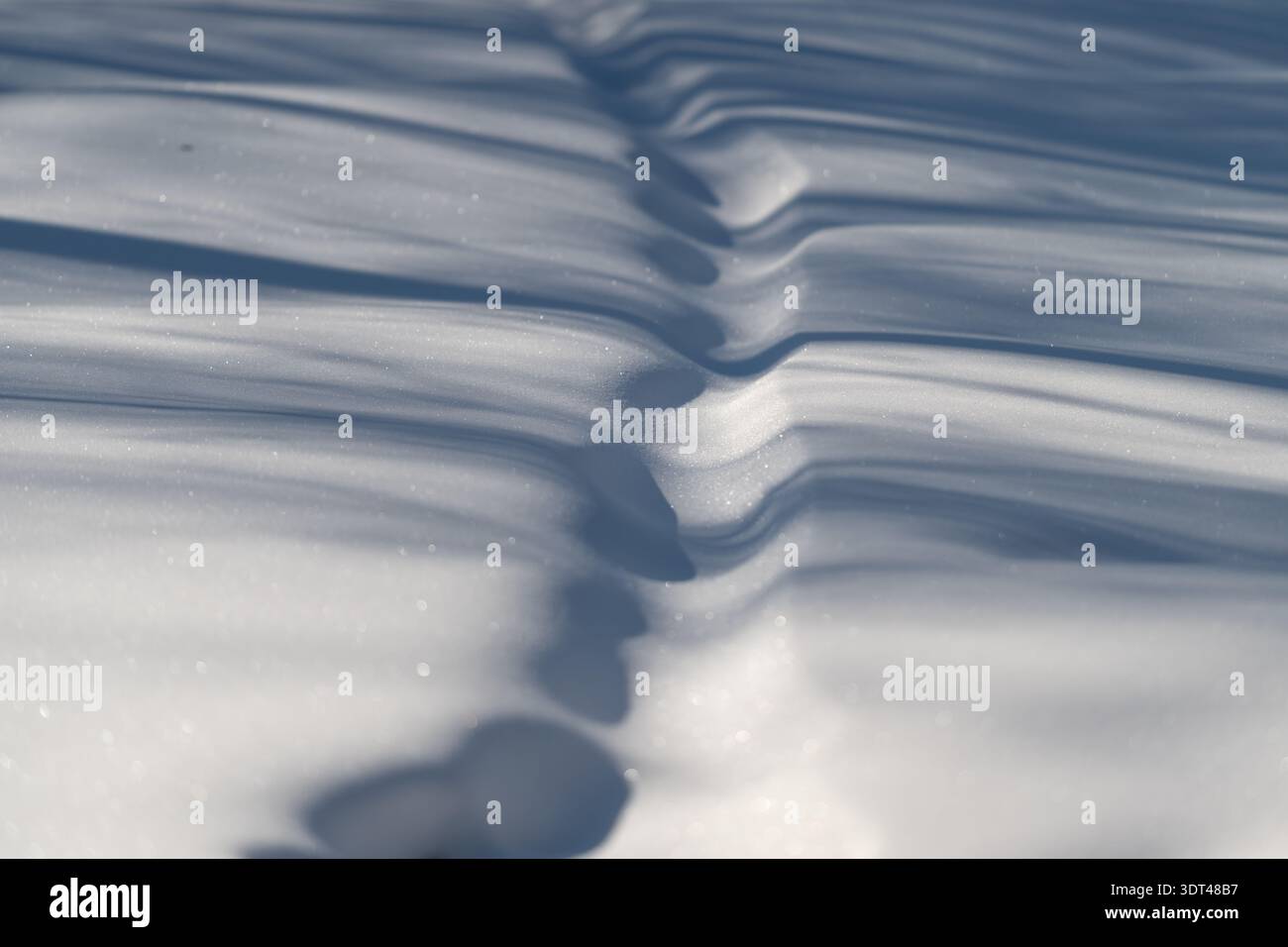 Frozen ripples form scenic waves. Repeated snow ripples generate undulating visuals with sunlight creating shadows and shimmering mountain tops Stock Photo