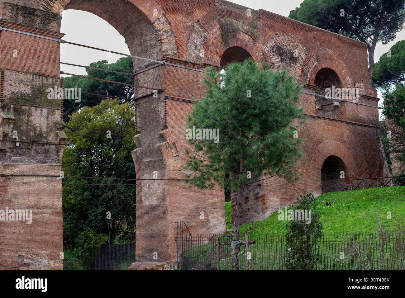 Rome, Italy – Brick arches of the ancient Aqua Claudia aqueduct rise above a grassy slope and pine trees, with metal supports and fencing visible in t Stock Photo
