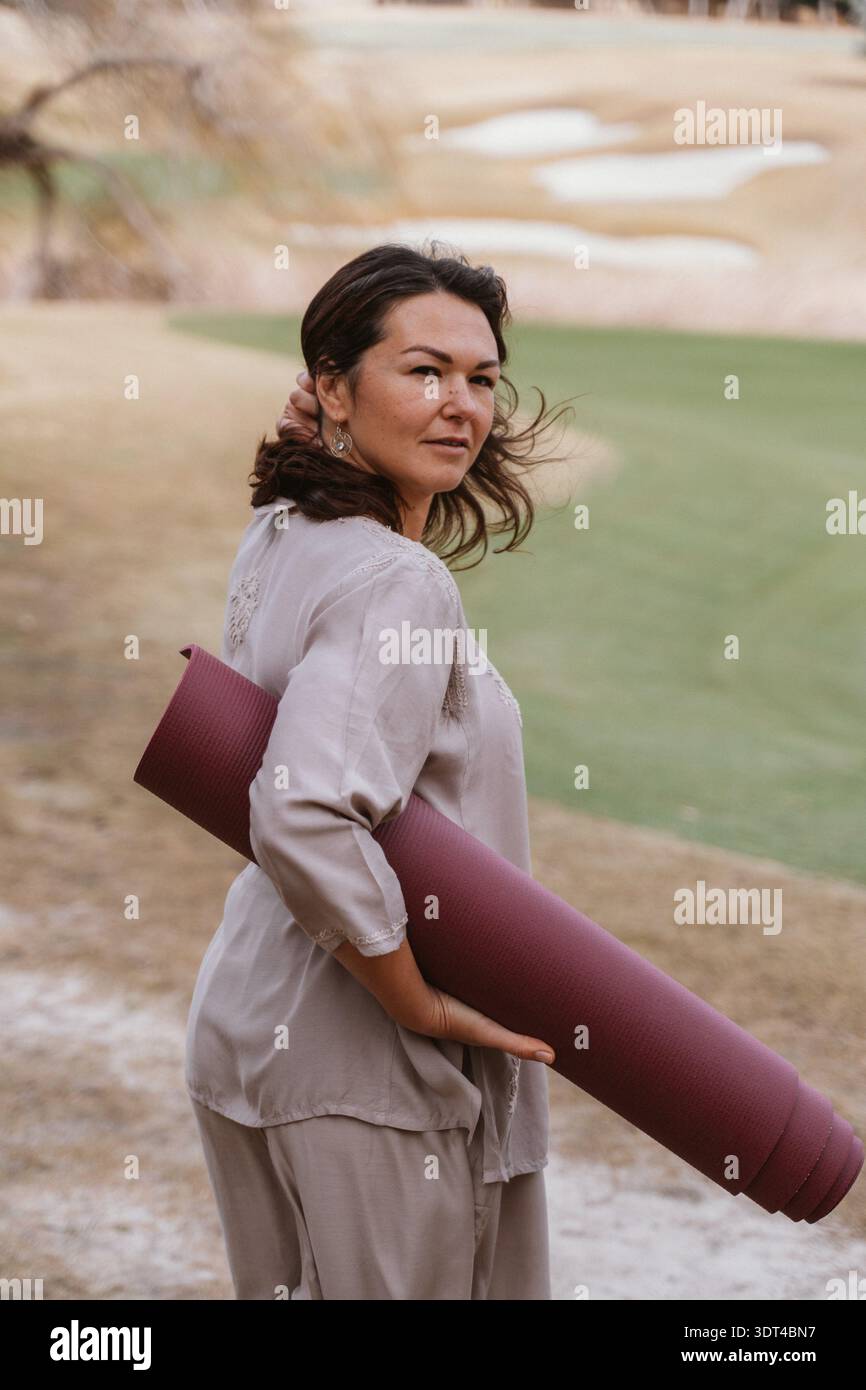 Woman standing outside holding a rolled yoga mat. Her expression is calm and focused, conveying tranquility. The outdoor setting adds a natural and se Stock Photo