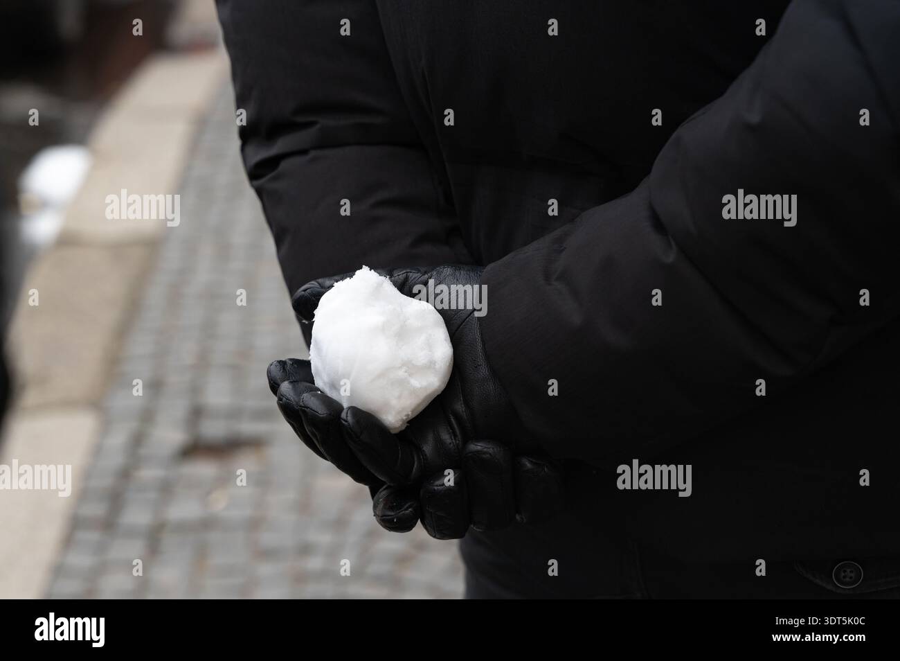 Hands holding snowball behind back, leather gloves clasping compact snowball, close crop on palms, textured cobblestone background, playful poised ges Stock Photo