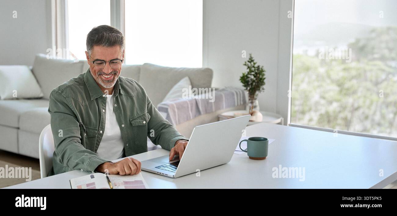 Happy older middle aged man using laptop computer at home table. Stock Photo