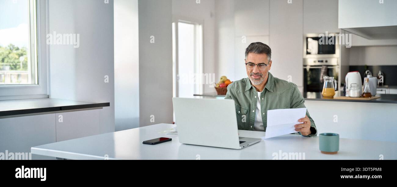 Senior mature man looking at laptop checking bill working at home kitchen table. Stock Photo