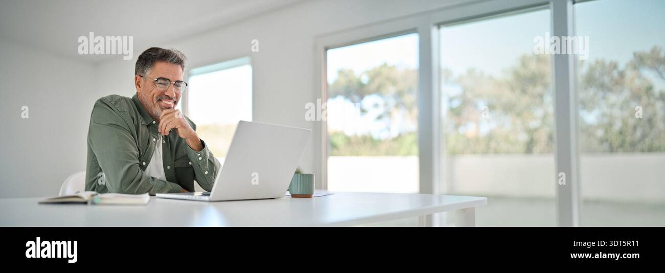 Happy older mature man using laptop computer at home table. Stock Photo