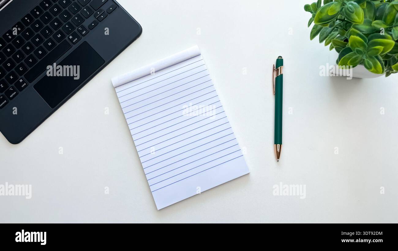 A blank notepad next to a pen, laptop, and houseplant on a white table. Concept for planning, ideas, notes, and workspace organization. Stock Photo