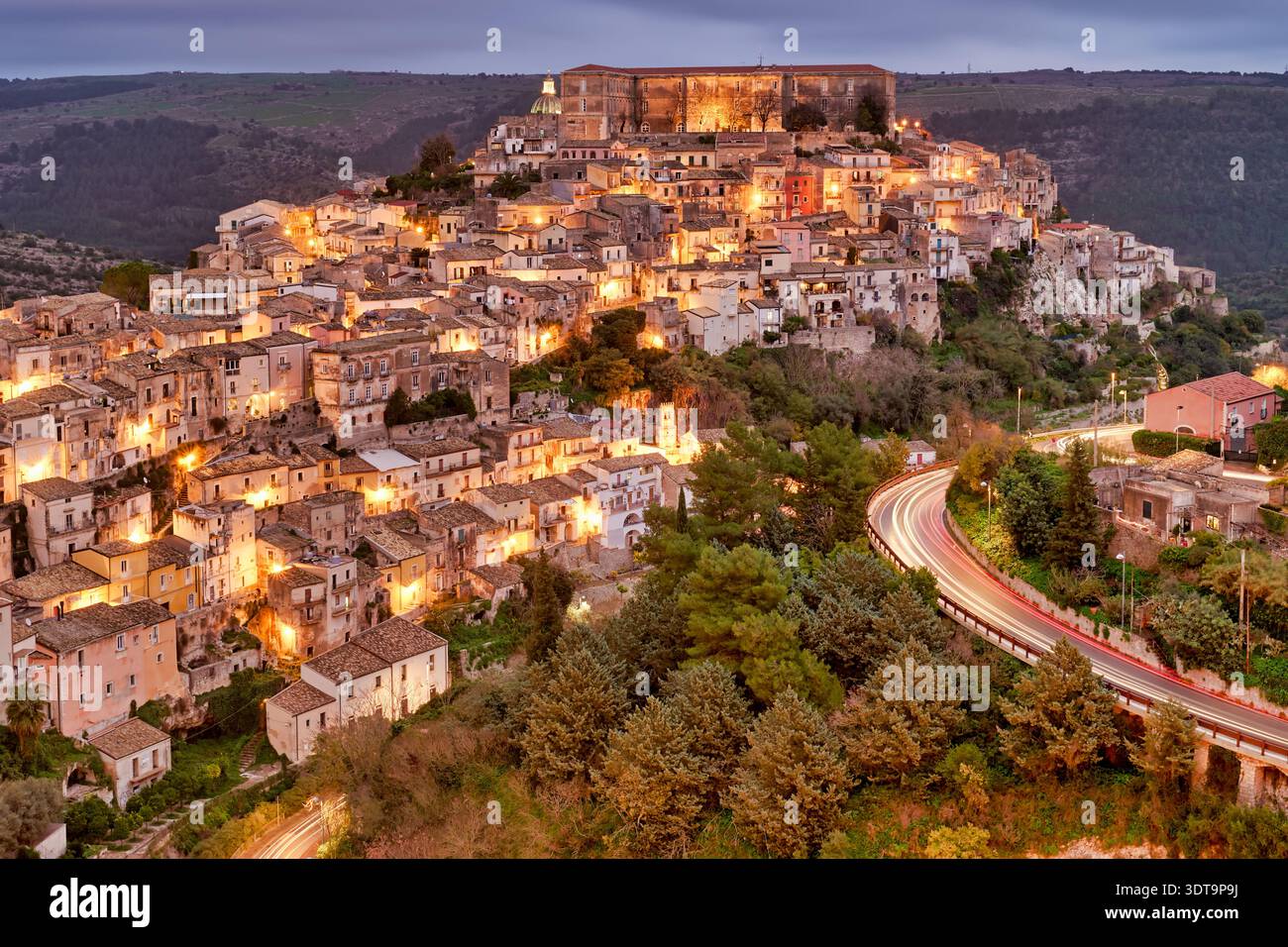 Ragusa Ibla old town shows buildings lit up at sunset with a winding road and hills around. This scene captures the beauty of Sicily. Stock Photo