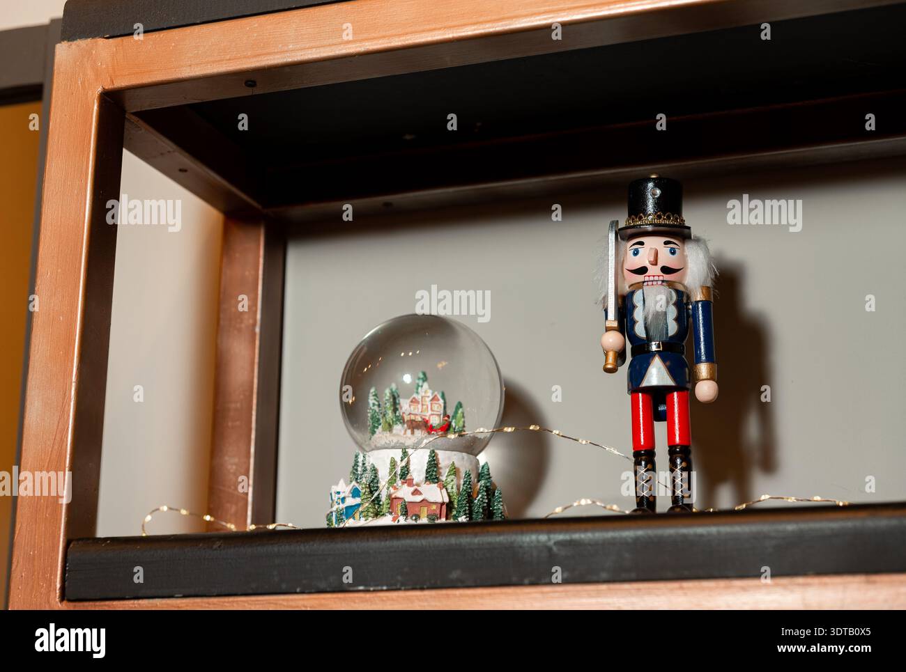 Festive holiday décor on a wooden shelf featuring a classic nutcracker figurine, a winter village snow globe and warm string lights against a neutral Stock Photo
