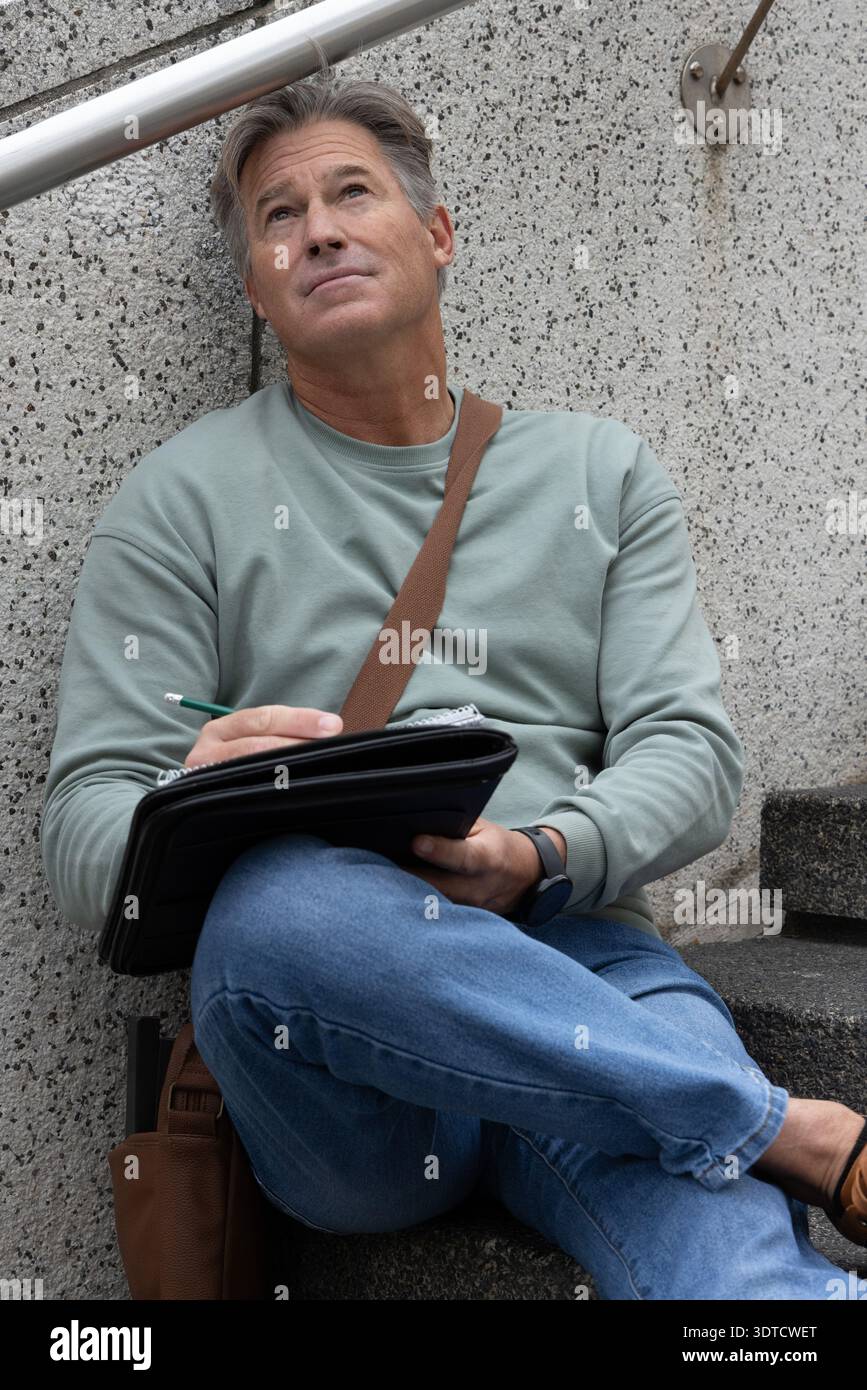 Middle-aged man sitting on concrete steps writing in black folio with green pencil Stock Photo