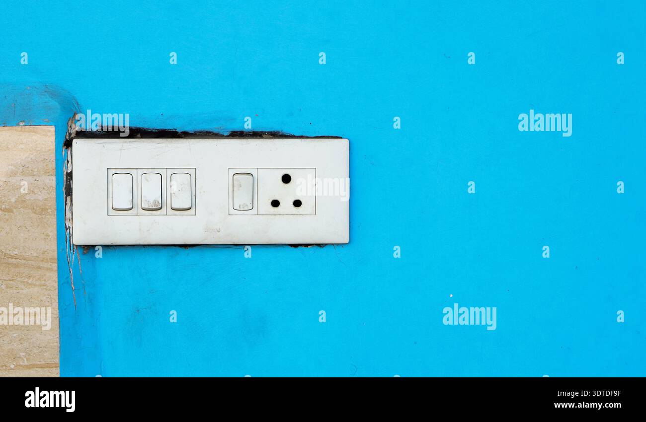 A white electrical switchboard with multiple switches and a socket installed on a bright blue wall. Stock Photo