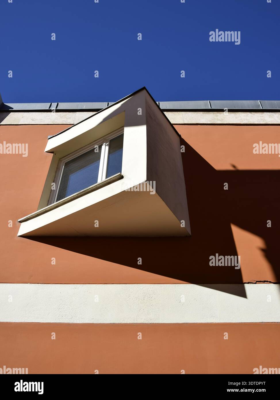 A protruding white-framed window casting a sharp angular shadow on a terracotta and beige striped wall. Stock Photo