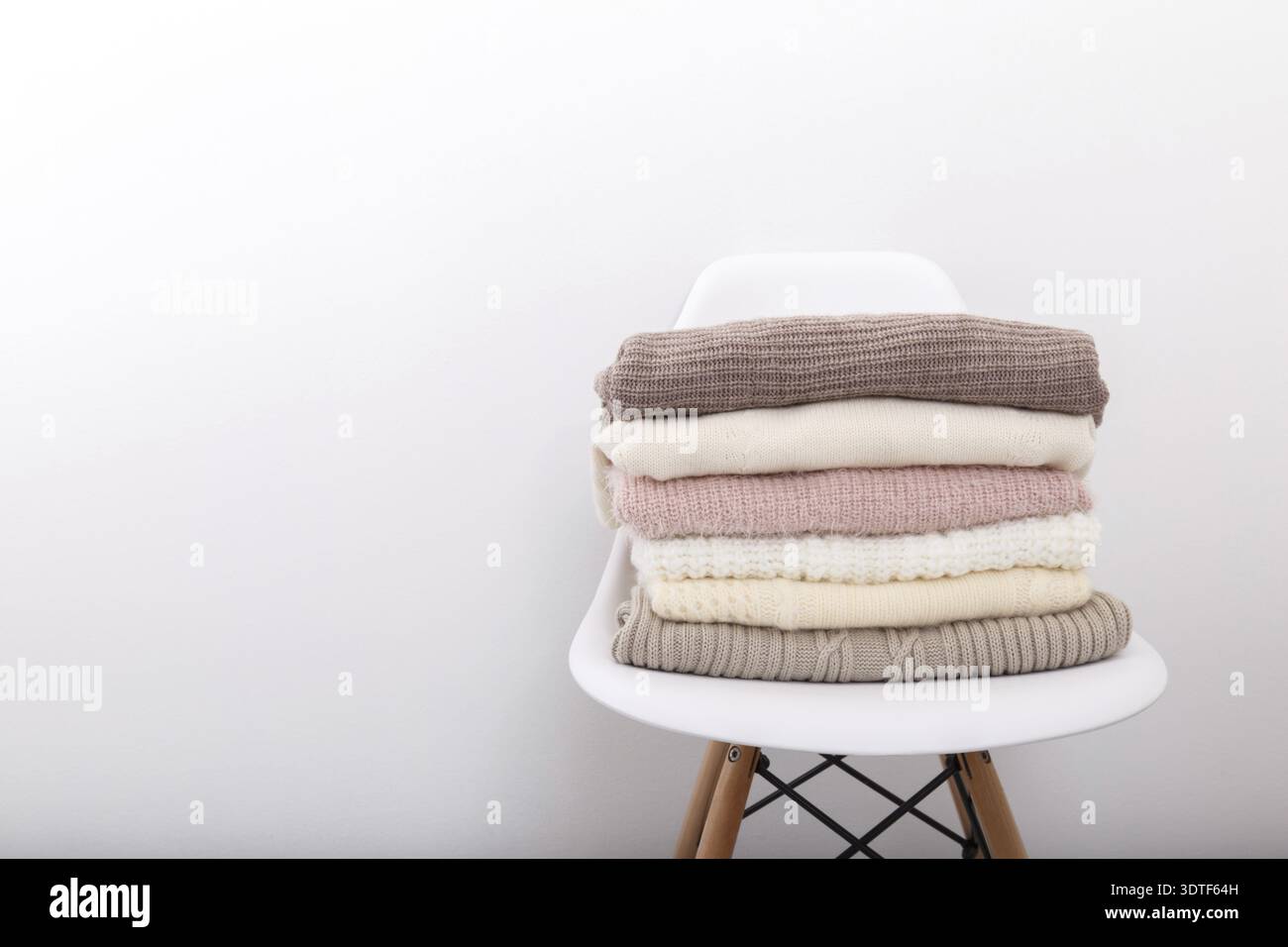 Stack of cozy knitted sweaters or blankets pastel trending colors on white modern chair against the wall Stock Photo