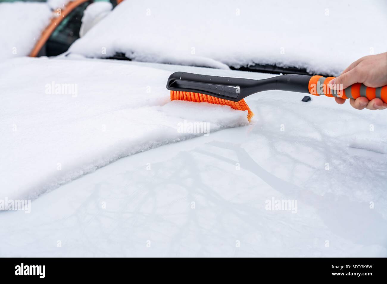 Snow clearing on car with orange brush in driveway. Family winter scene with snow removal using telescopic brush in driveway Stock Photo