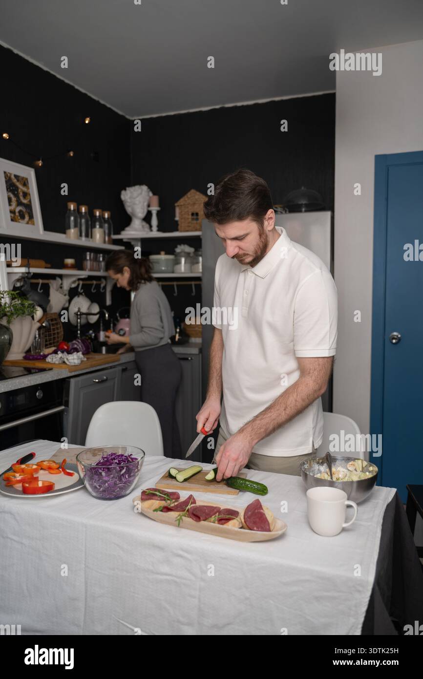 A couple is seen actively preparing a meal in a cozy kitchen. The man slices cucumbers while the woman focuses on tasks by the counter, surrounded by Stock Photo