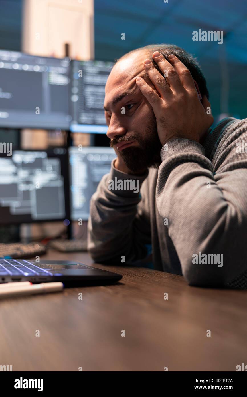 Overworked programmer rests head in palms during difficult coding session. Male developer analyzes complex data on laptop with exhausted expression, represents programming stress and technical pressure Stock Photo