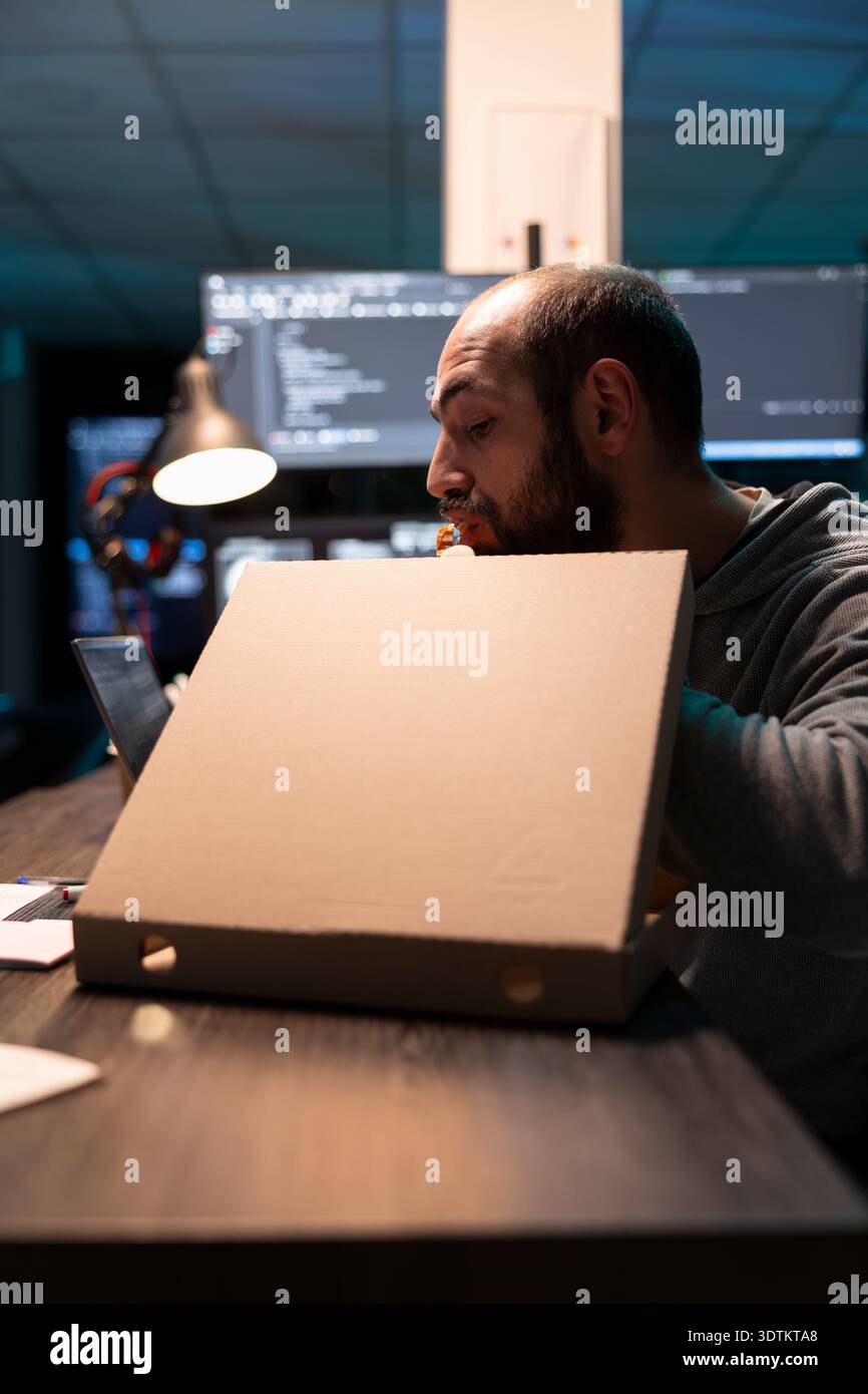 Software developer eating pizza straight from box and analyzing decoding information on laptop. Screens filled with code surrounds programmer, representing busy work routine and late hour productivity Stock Photo