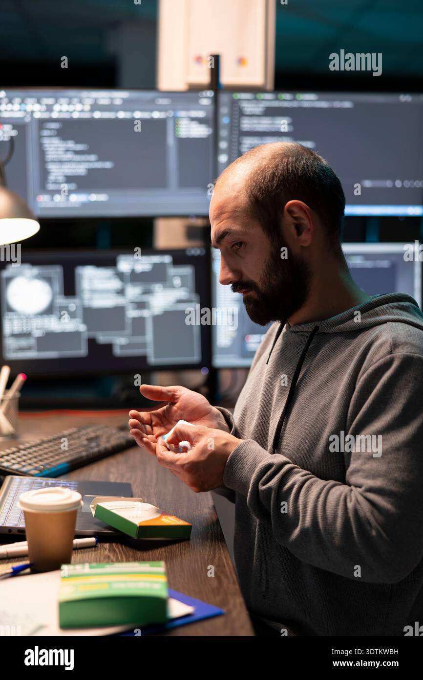 Male IT worker holds medication at desk, managing fatigue after long hours solving technical problems. Caucasian programmer checks pills while surrounded by monitors displaying complex system code. Stock Photo