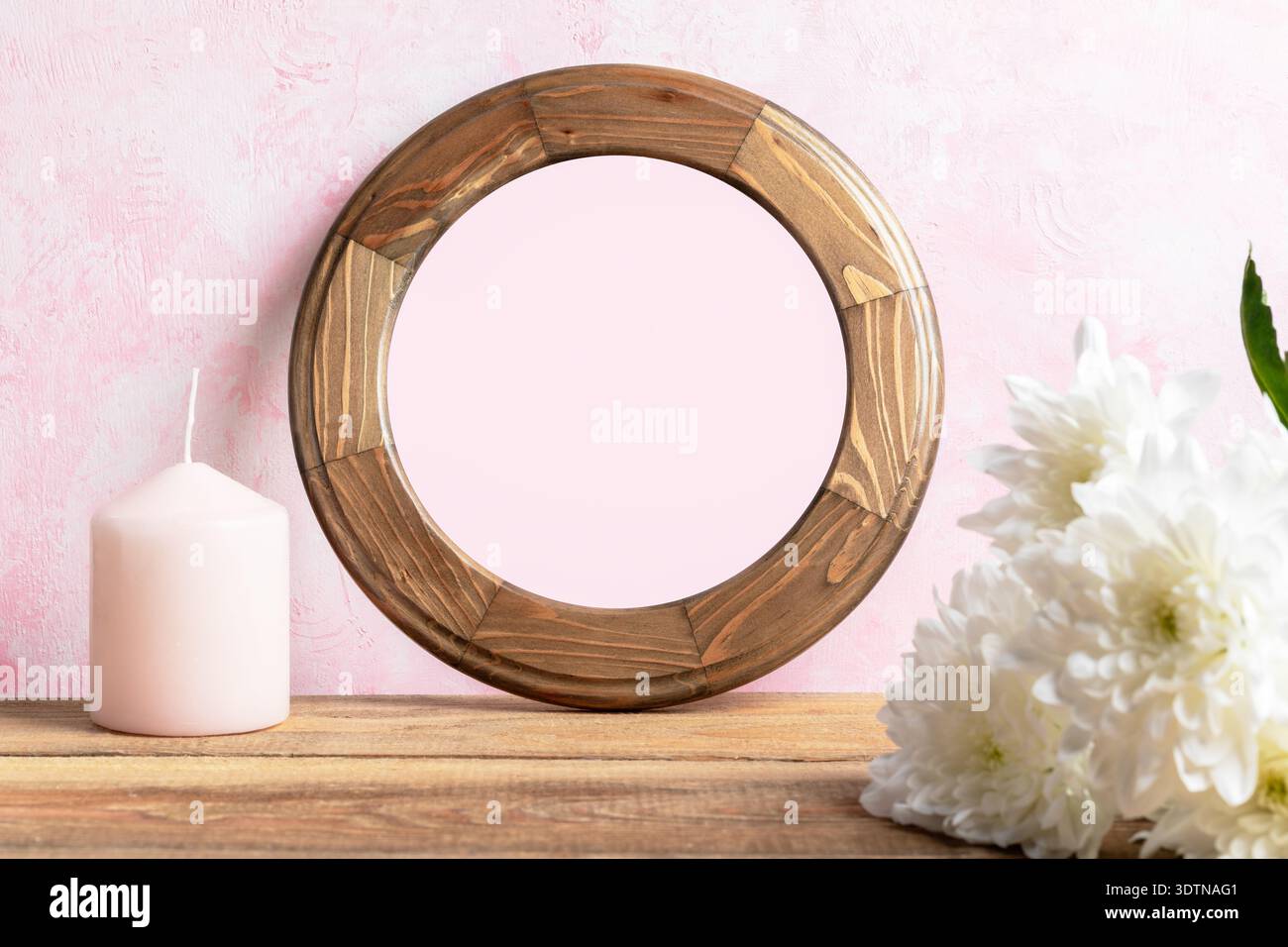 Round blank wooden frame with natural finish rests on wooden table beside pink candle and white flowers set against soft pink wall, creating beautiful Stock Photo