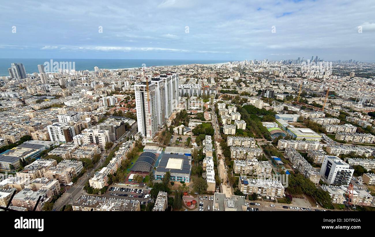 High-angle aerial view of a dense coastal city featuring modern skyscrapers, residential blocks, and a sports arena stretching toward the blue sea. - Smartphone Captured Stock Image