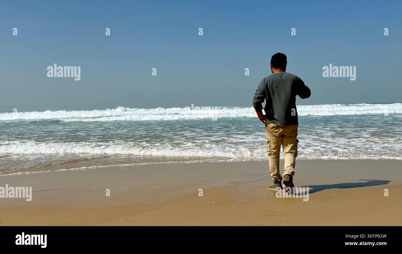 Rear view of a man in cargo pants walking on a sunny beach towards crashing ocean waves. A peaceful scene of solitude, travel, and coastal nature. - Smartphone Captured Stock Image