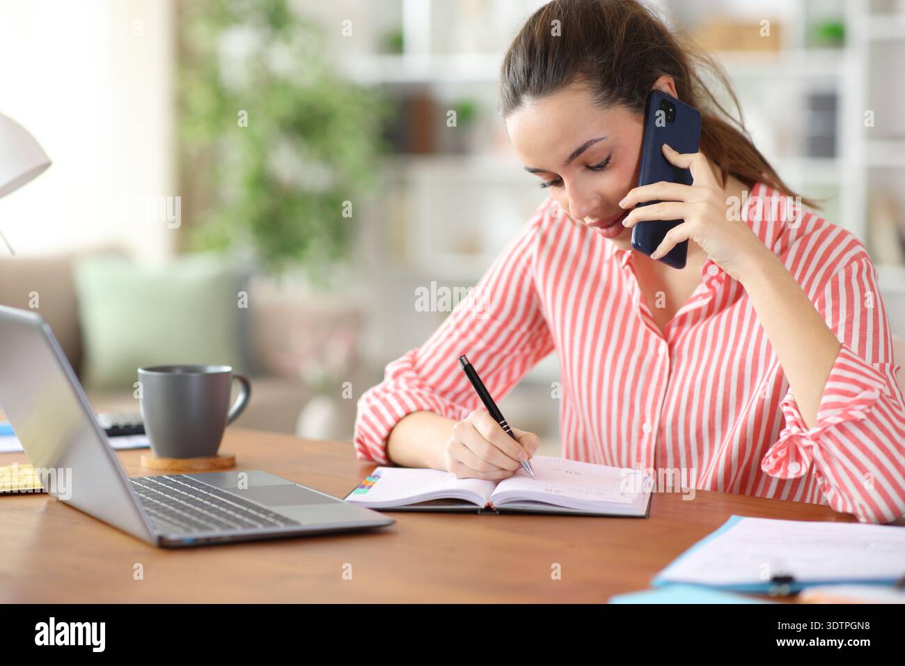 Freelance worker writing in agenda talking on phone working at home Stock Photo