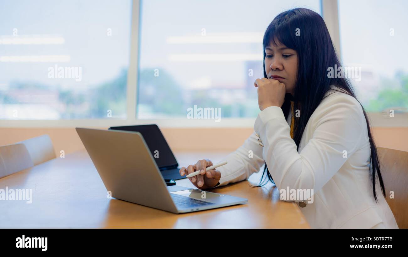Asian businesswoman working on laptop and tablet in office. Female professional thinking, analyzing data, using stylus pen for digital tasks. Professi Stock Photo