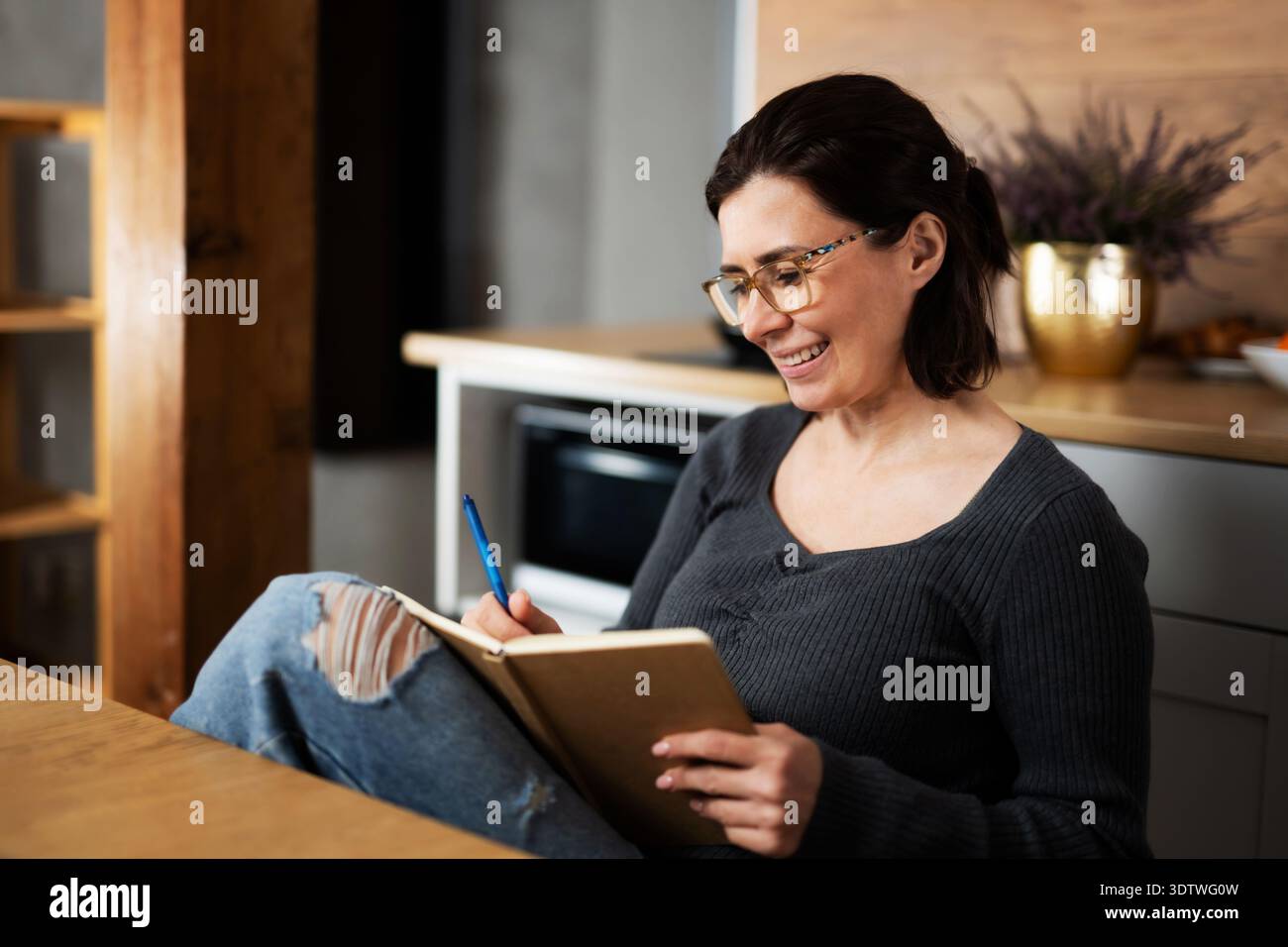 Happy Young Woman Writing Notes In A Notepad In The Kitchen Stock Photo