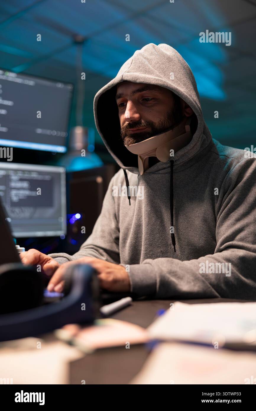 Hooded injured man working diligently on cybersecurity, analyzing networks and spyware risks. Focused male programmer with neck brace writing code and concentrating on improving company networks. Stock Photo