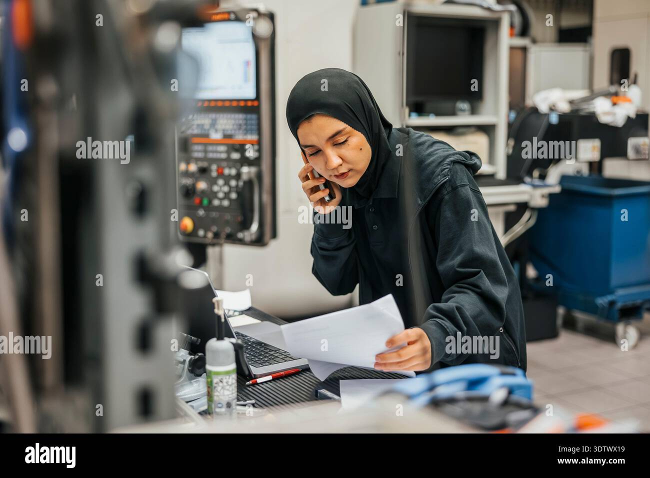 Young female employee talking on phone while reading document in metal factory Stock Photo