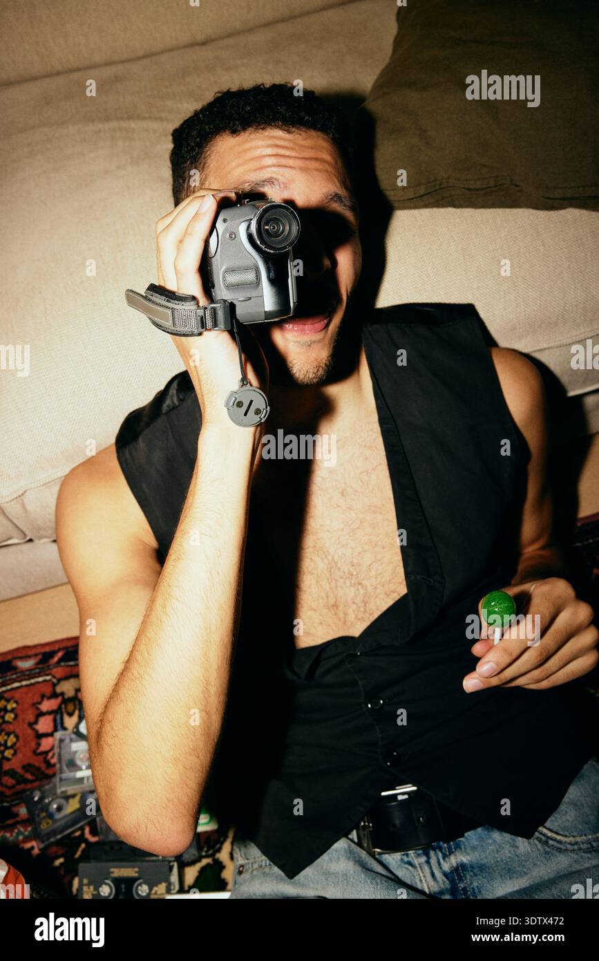 Young man holding candy while looking through vintage video camera at home Stock Photo