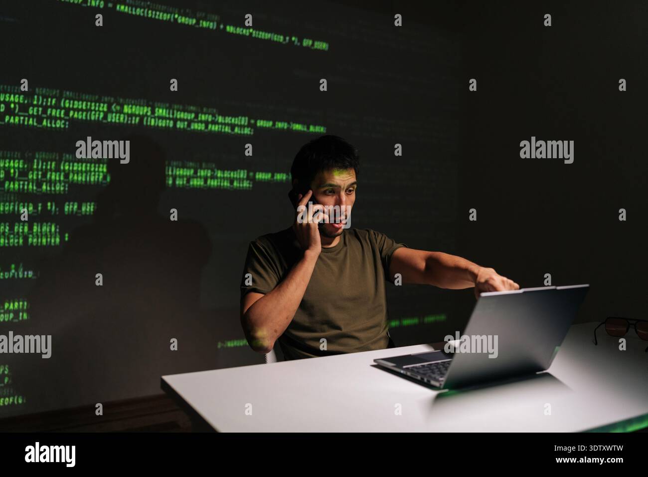 Busy programmer male sitting at desk with laptop, deeply engaged in phone conversation while analyzing projected green code, symbolizing complex Stock Photo