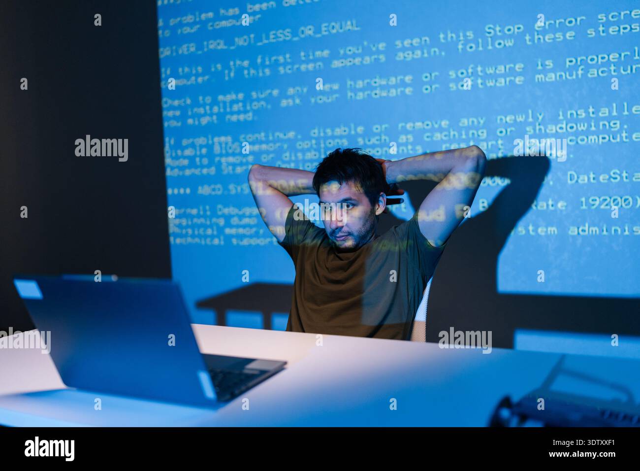 Thoughtful young male developer focused on laptop, debugging code late at night as blue light and projected code lines create cybersecurity and Stock Photo