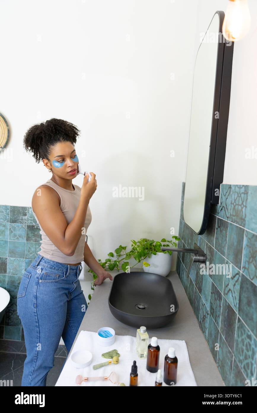 African American woman using skincare, wearing under-eye patches at bathroom sink, copy space Stock Photo