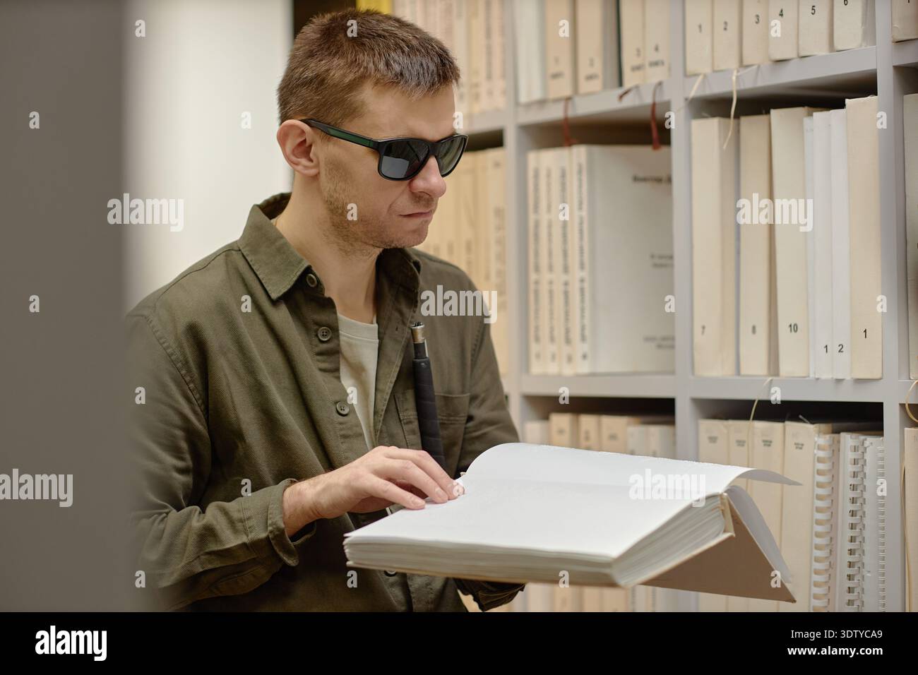Young Man Reading Braille Book in Library While Using Cane Stock Photo