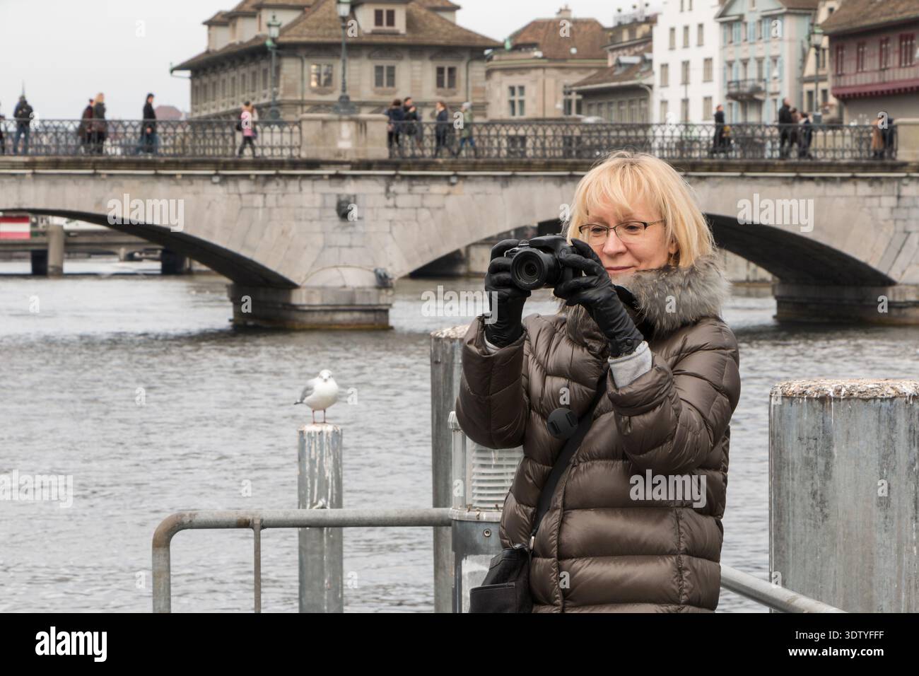 Blonde attractive woman in modern glasses making photo on camera against city bridge in cloudy winter day. Europe cityscape. Stock Photo