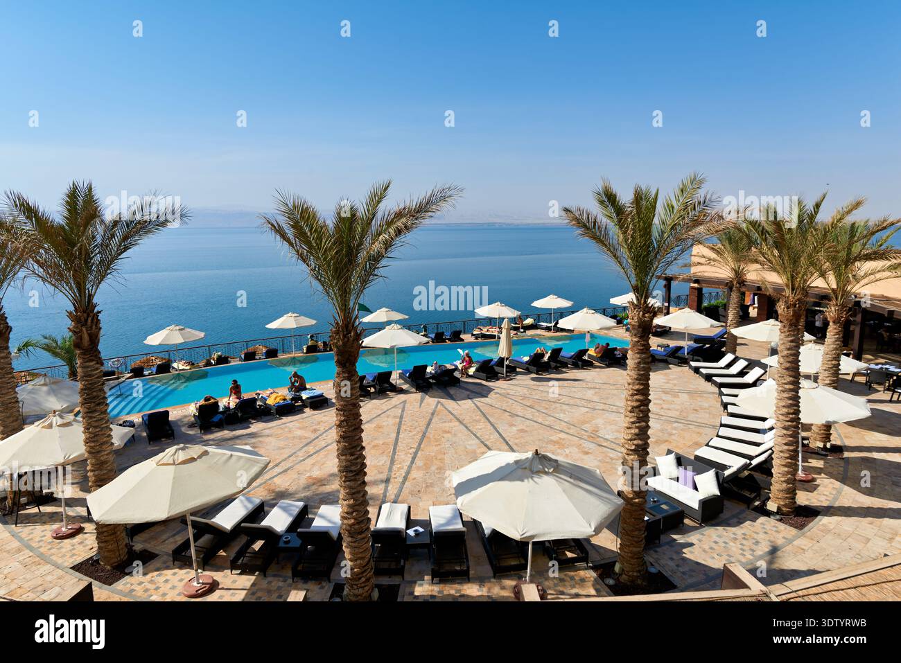 People enjoy the pool area at Movenpick Resort, overlooking the Dead Sea. Guests relax under umbrellas on a sunny day by the water. Stock Photo