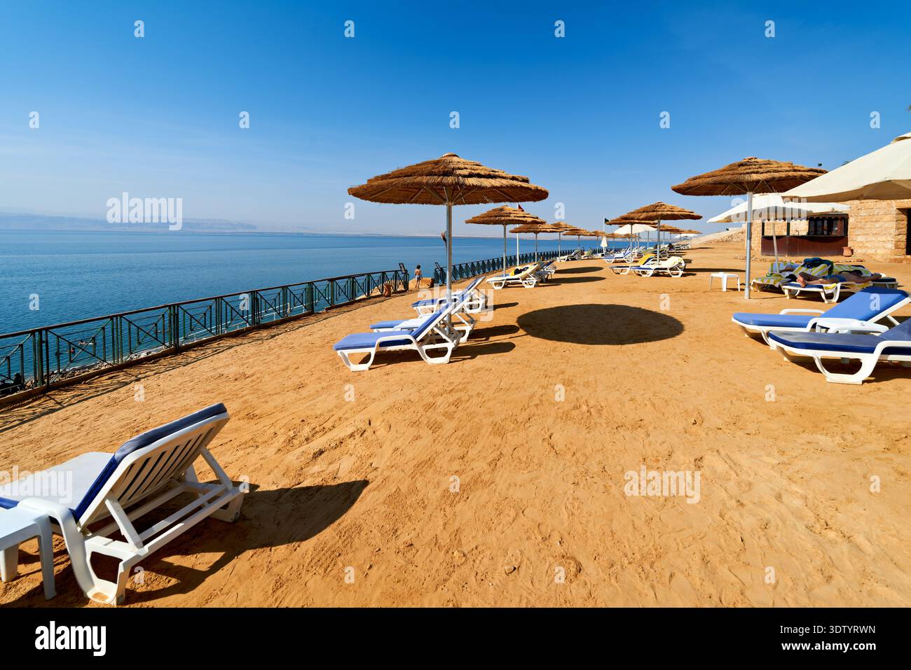 Visitors enjoy lounge chairs at Movenpick Resort, facing the Dead Sea. Sunlight shines on the sandy beach and calm water. Stock Photo