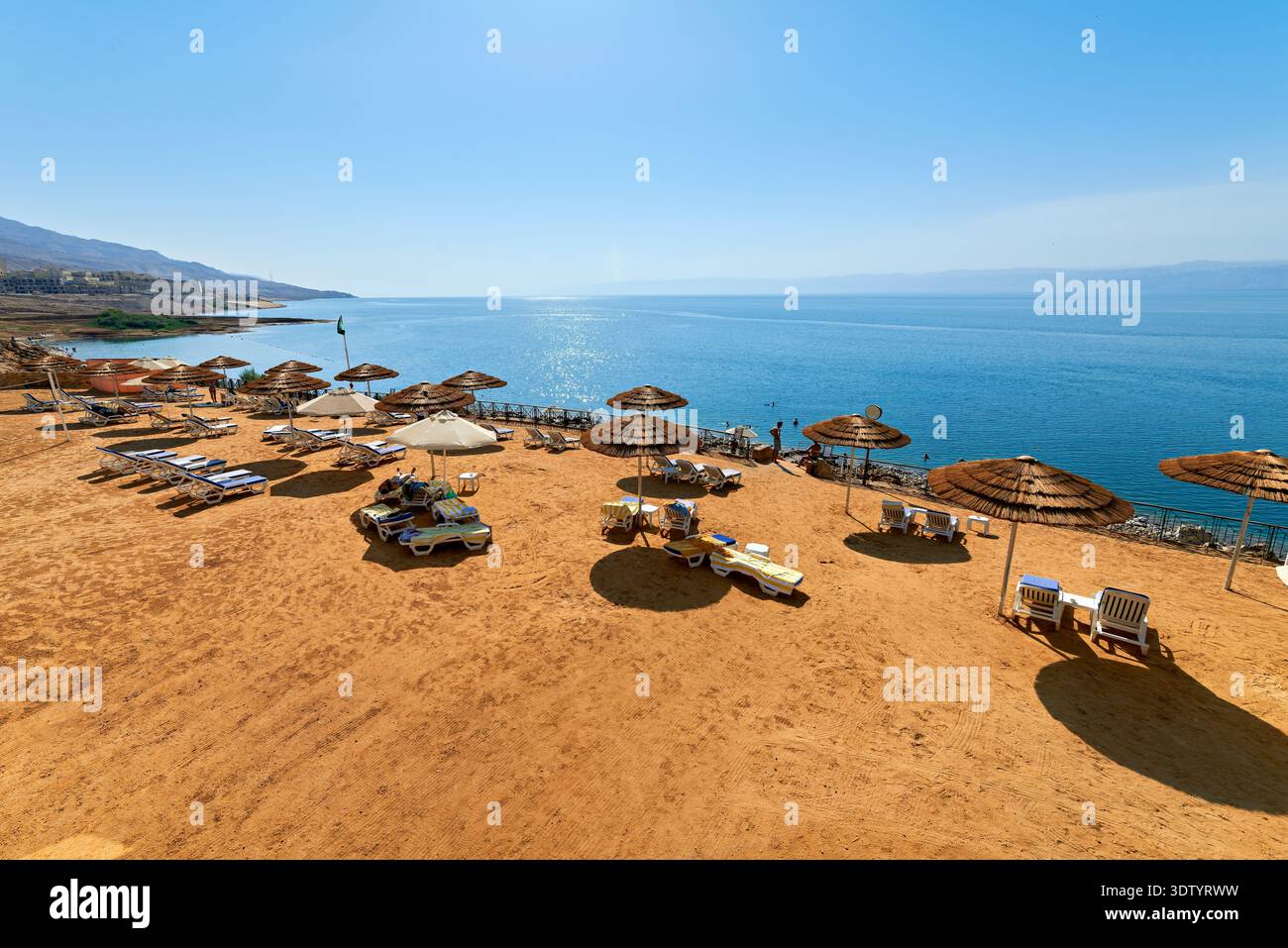 Visitors relax by the beach at Movenpick Resort at the Dead Sea in Jordan. Sunbeds and umbrellas are set up on the sand facing the water. Stock Photo