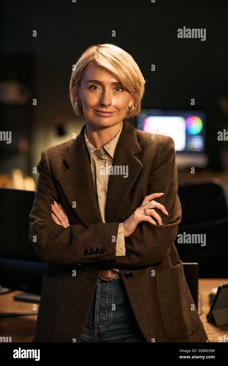 Portrait of middle aged Caucasian woman standing with arms crossed, smiling confidently while looking into camera, office environment with computer monitor in background visible Stock Photo