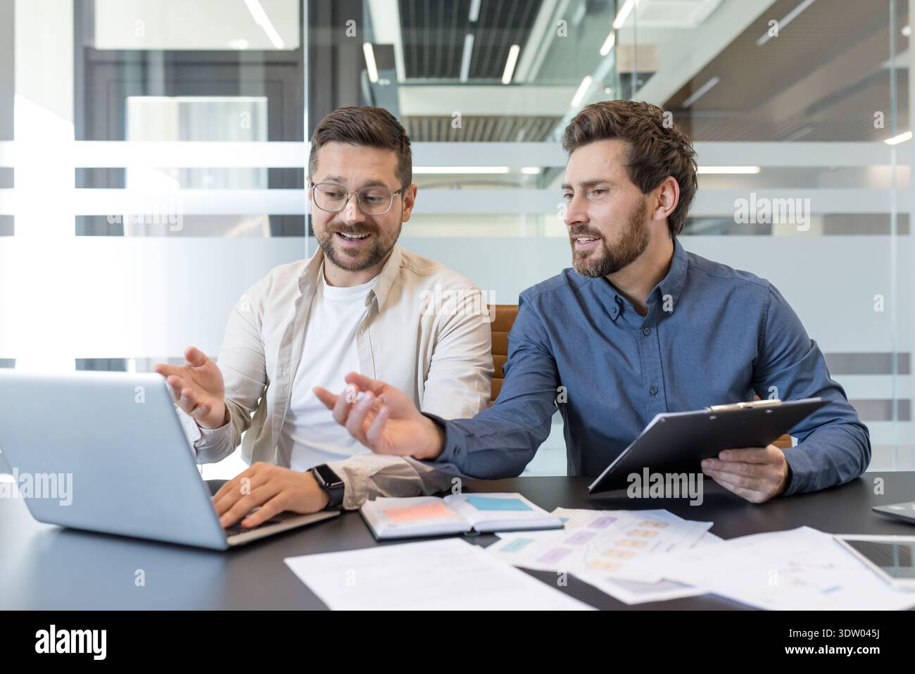 Two businessmen discussing project details and ideas while working together on a laptop, fostering teamwork and professional collaboration in a modern office environment Stock Photo