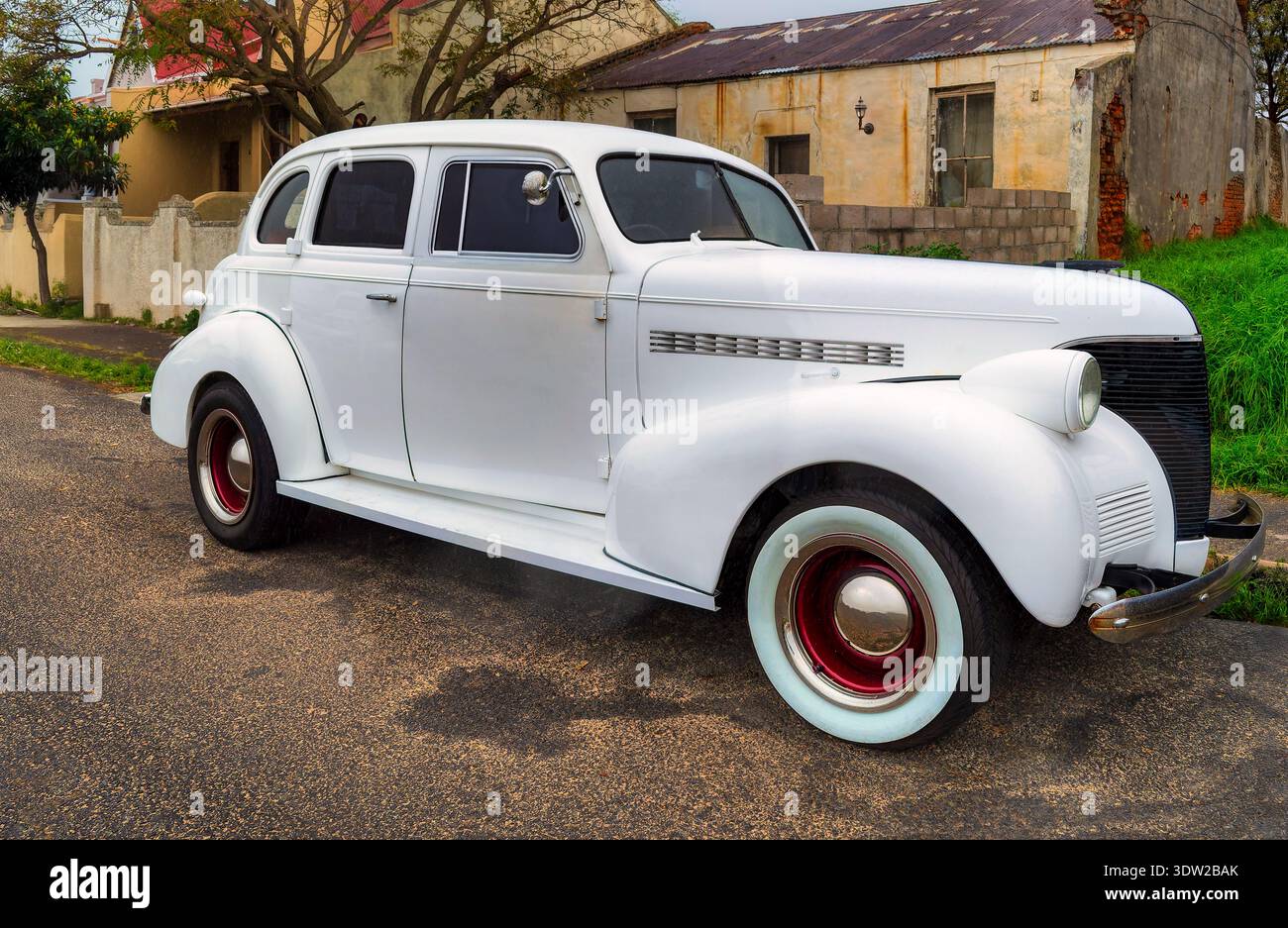 side profile view of a classic white four-door sedan from the late 1930s with whitewall tires and red rims, parked on a paved residential street in So Stock Photo