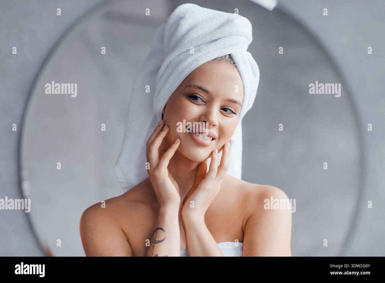 Woman With Towel on Head Smiles While Touching Her Face in a Bathroom Setting Stock Photo