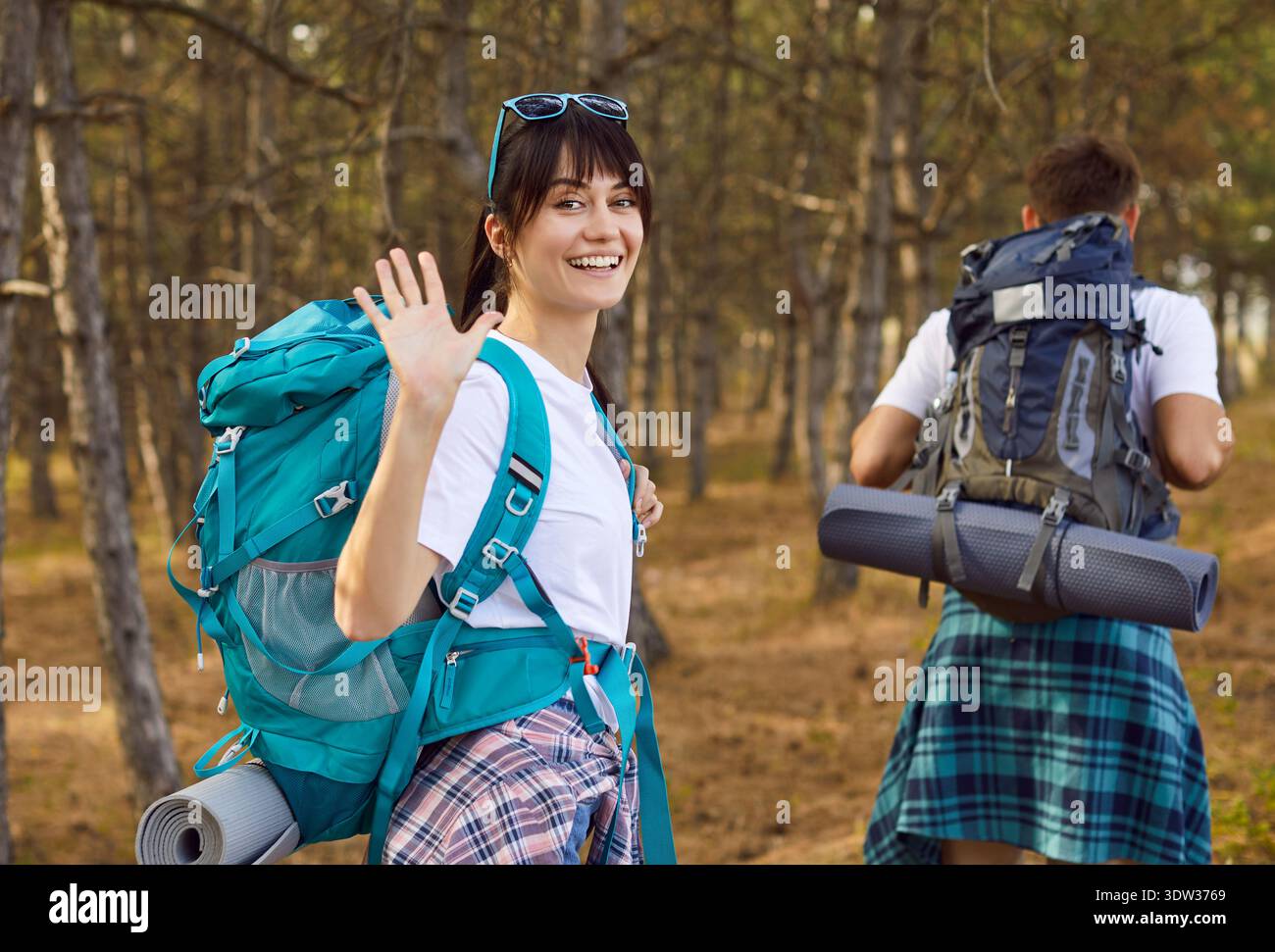 Smiling female hiker with backpack waving while partner walking on forest trail Stock Photo