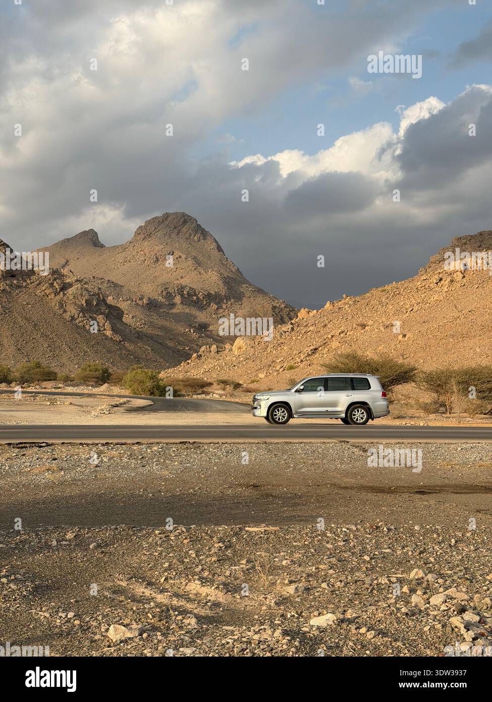 SUV driving along a remote desert road in the rugged Hajar Mountains near Muscat, Oman, with dramatic rocky landscape and cloudy sky in evening light - Smartphone Captured Stock Image