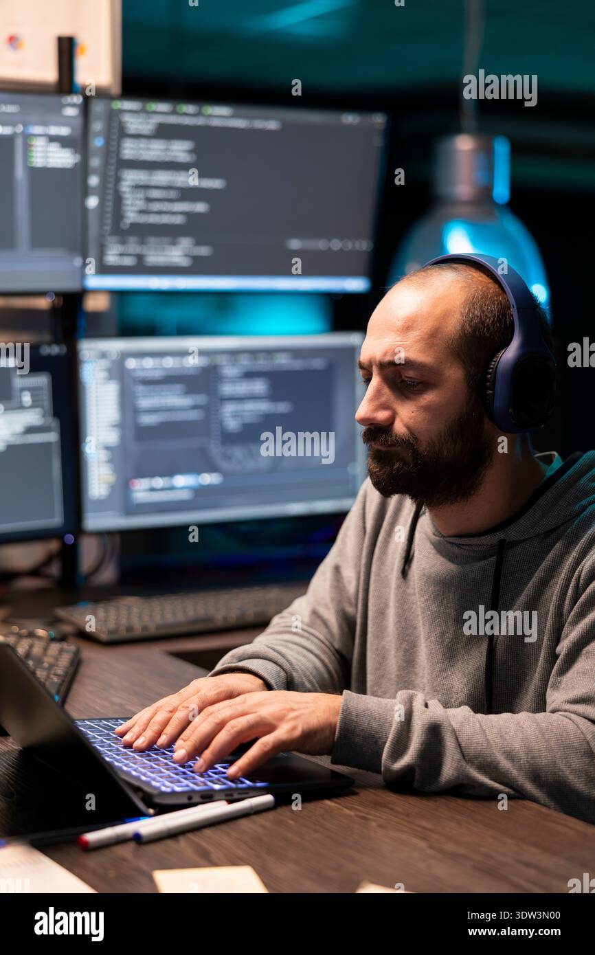 Male adult freelancer types on laptop in tech workspace, creating software while planning and debugging encrypted programs. White man works night hours on cybersecurity, analyzing spyware risks. Stock Photo