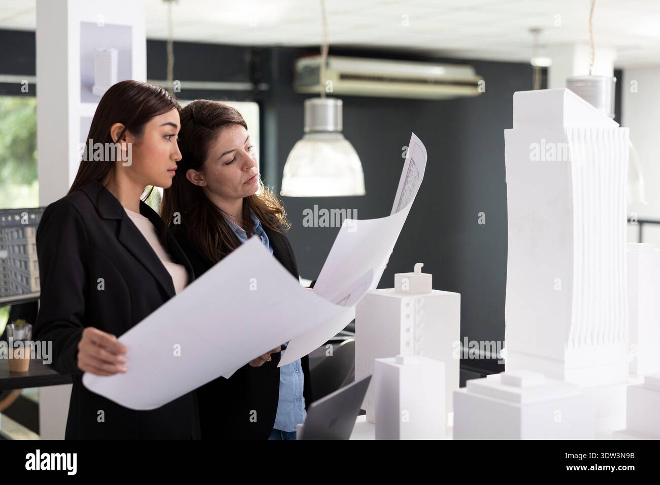 Skilled architects study printed architectural blueprints, preparing for next construction site evaluation. Professional women crosschecking design variations for accurate project implementation. Stock Photo