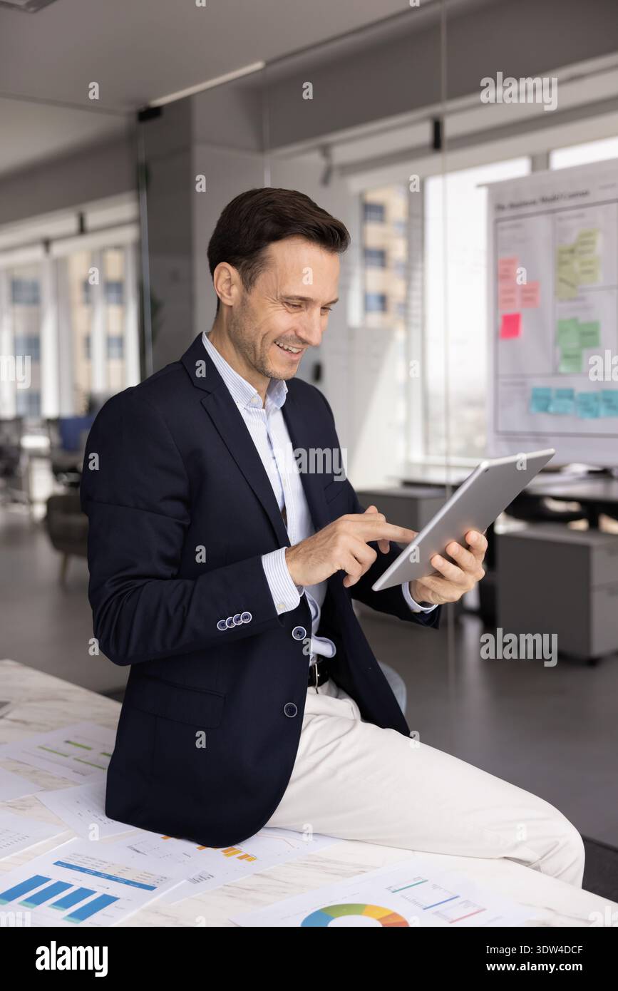 Smiling confident millennial businessman sitting on table using tablet device Stock Photo