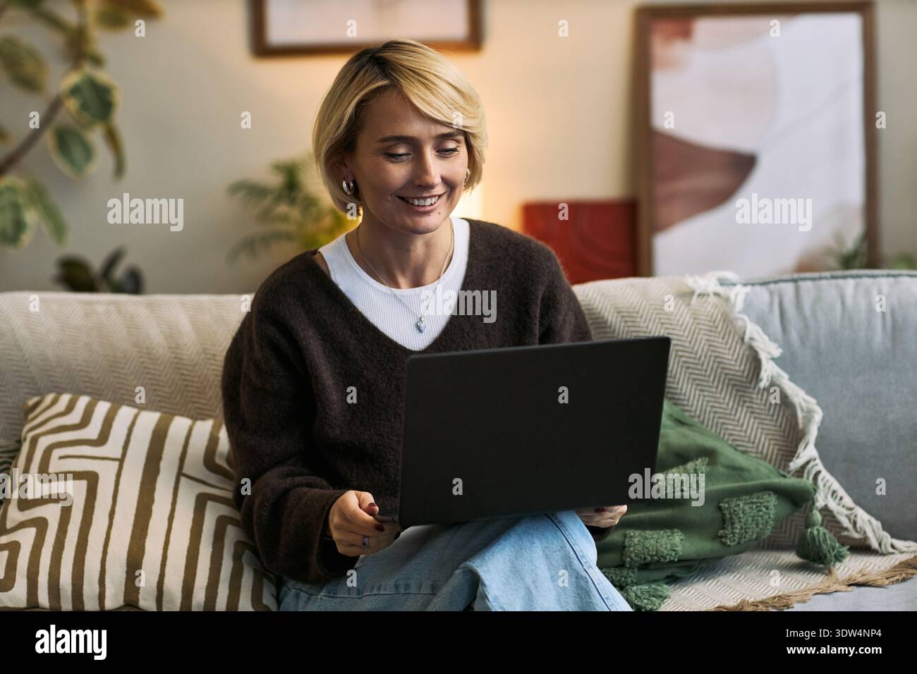 Caucasian middle aged woman sitting on sofa using laptop smiling while engaging with femtech health platform in home setting, demonstrating technology use for womens health management Stock Photo