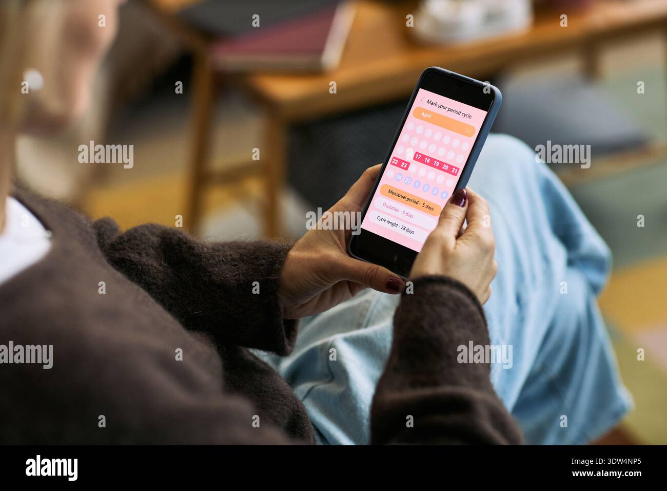 Caucasian woman holding smartphone using femtech health app for period tracking, sitting indoors focusing on menstrual cycle calendar interface on screen Stock Photo
