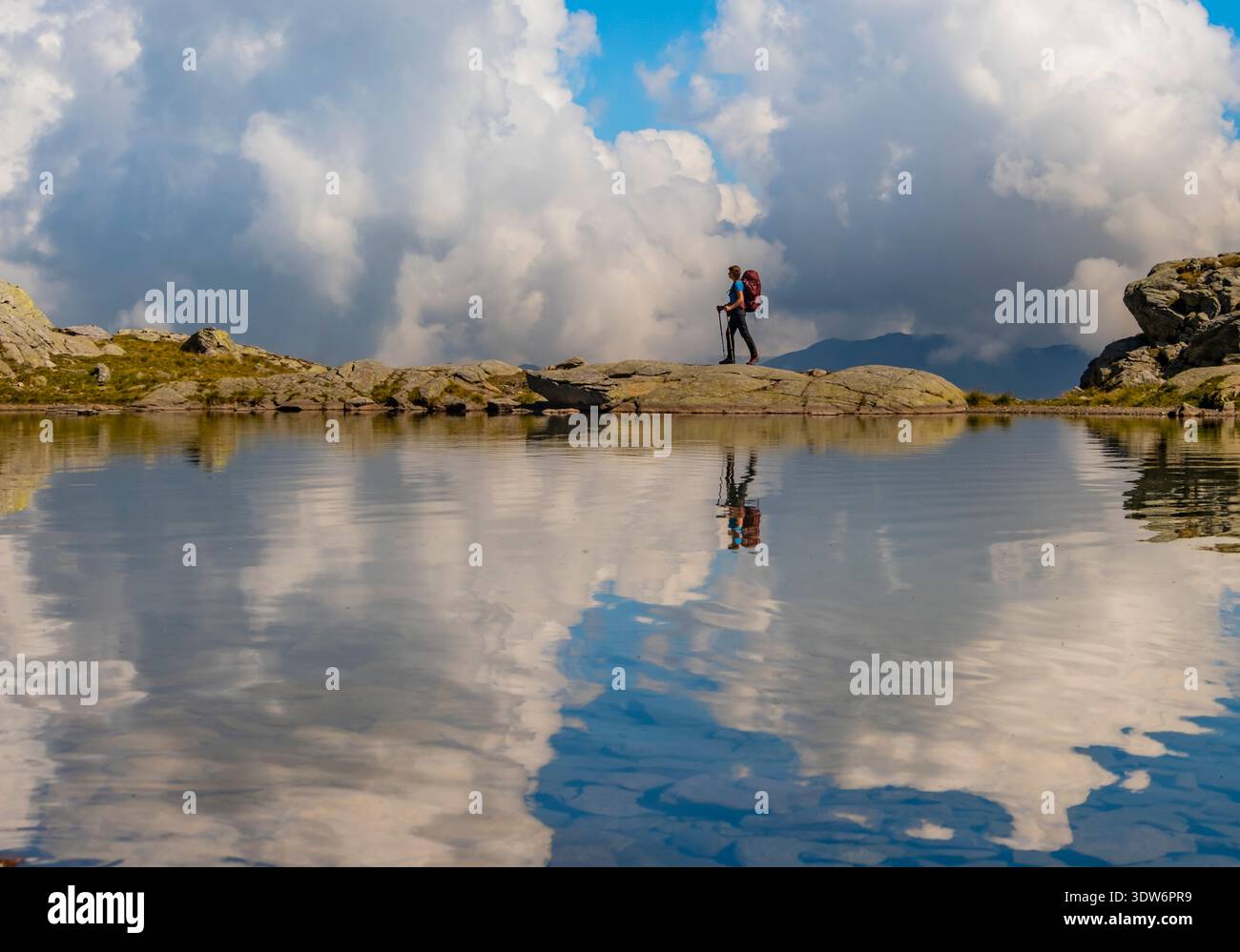 A hiker explores a serene alpine landscape in Swizz, surrounded by towering clouds and a calm lake. The reflection creates a picturesque scene of Stock Photo
