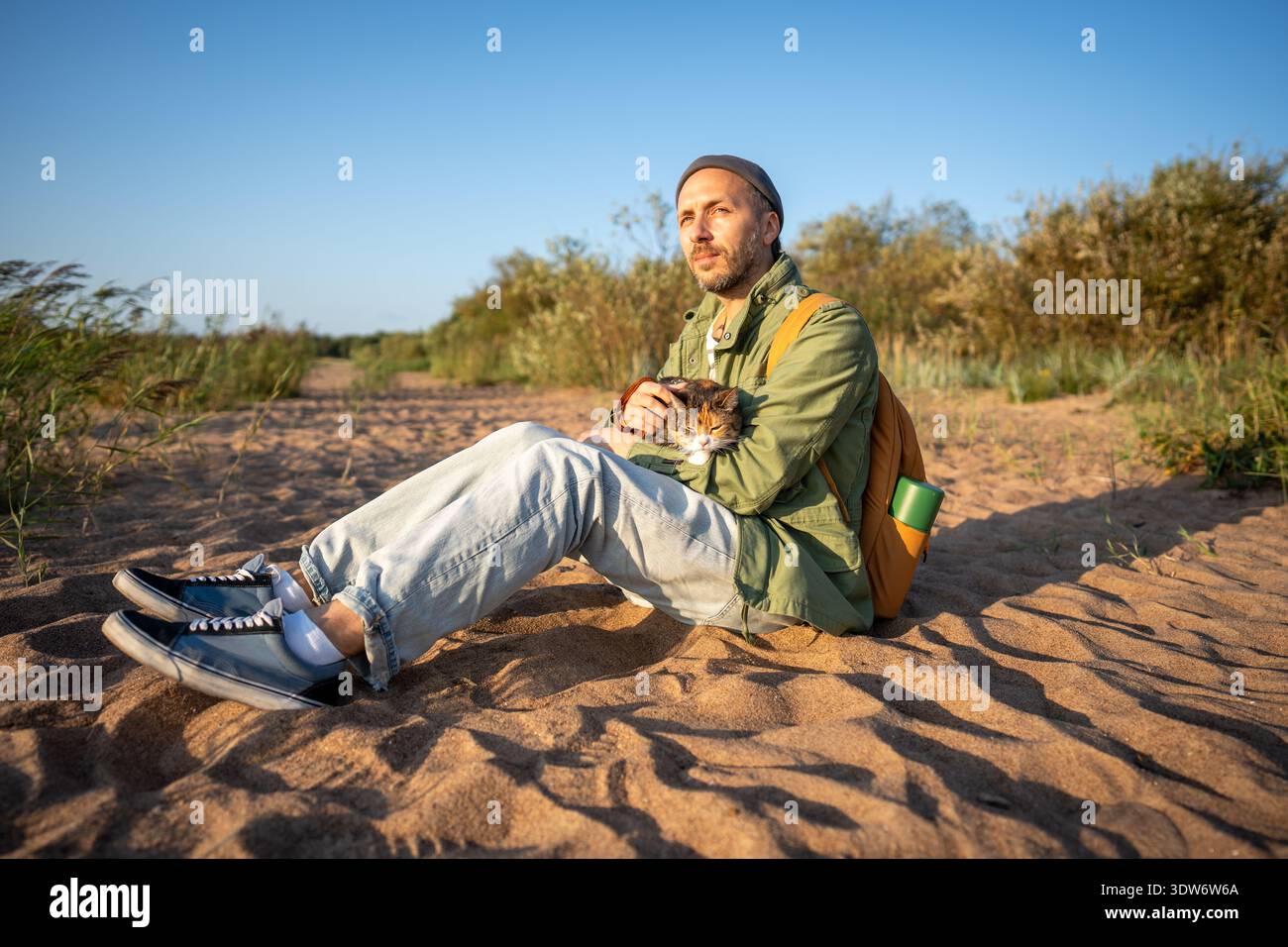 Thoughtful relaxed man rest on sandy beach holding cat in hands while enjoy fresh breeze together  Stock Photo
