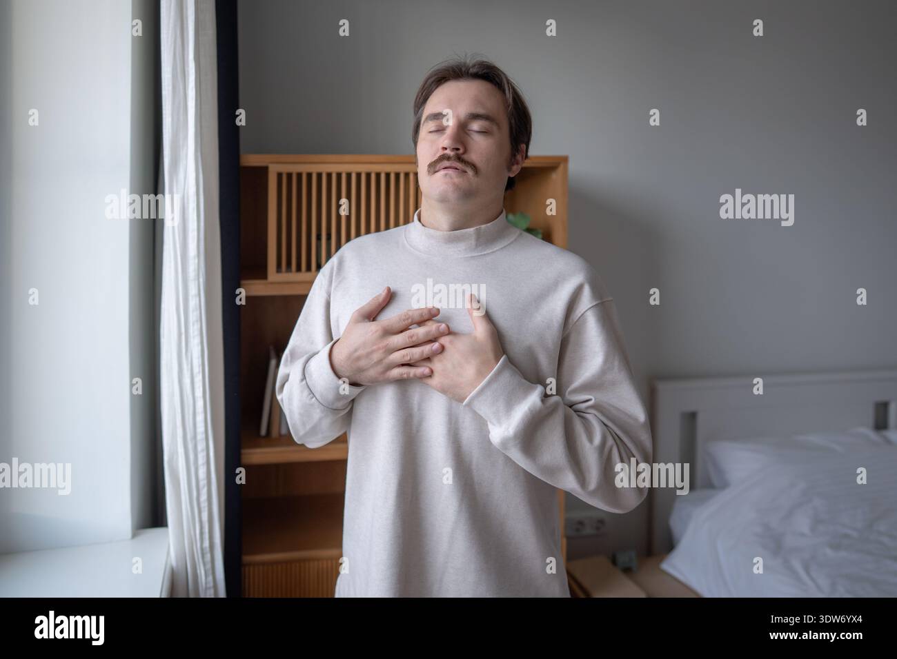 Calm reflective man with hands on chest, practicing deep breathing to ease emotional tension at home Stock Photo